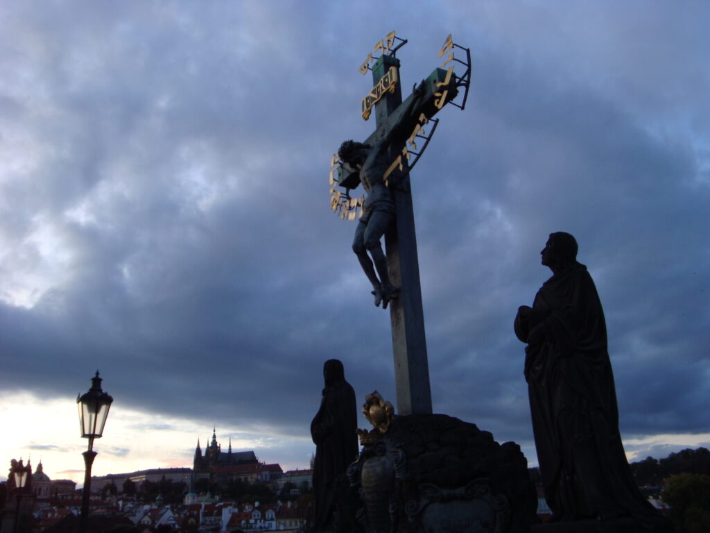 A cross statue at dusk with looming clouds in the background in Prague