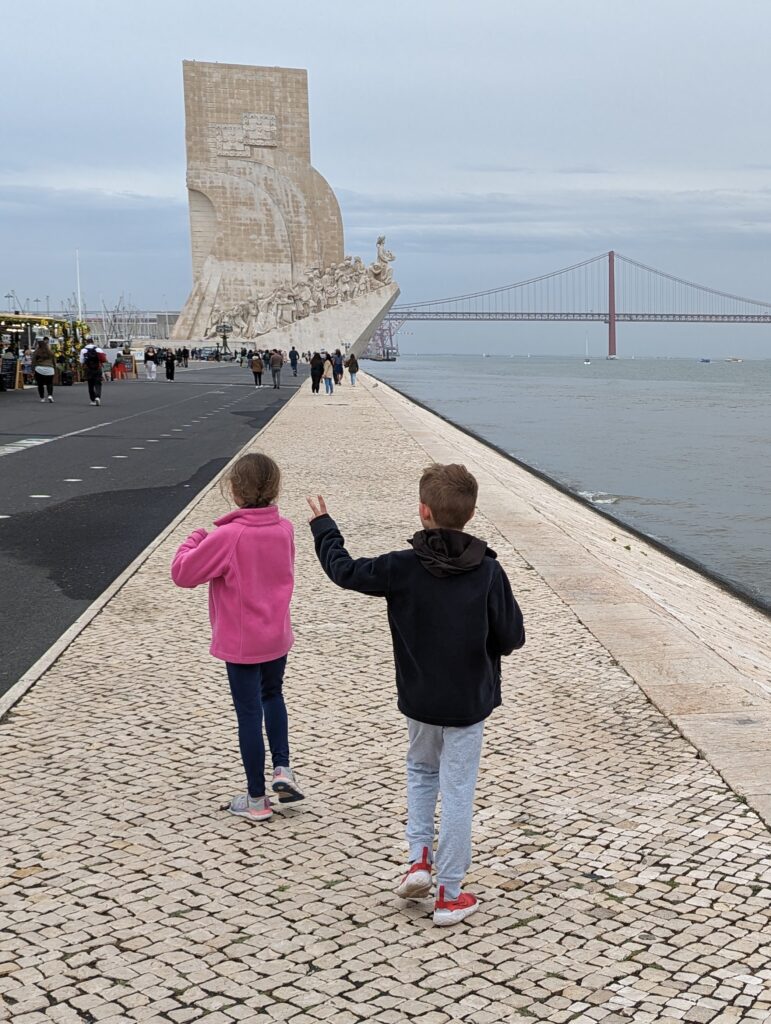 Two kids walking alongside water with a large monument and bridge in the background in Lisbon