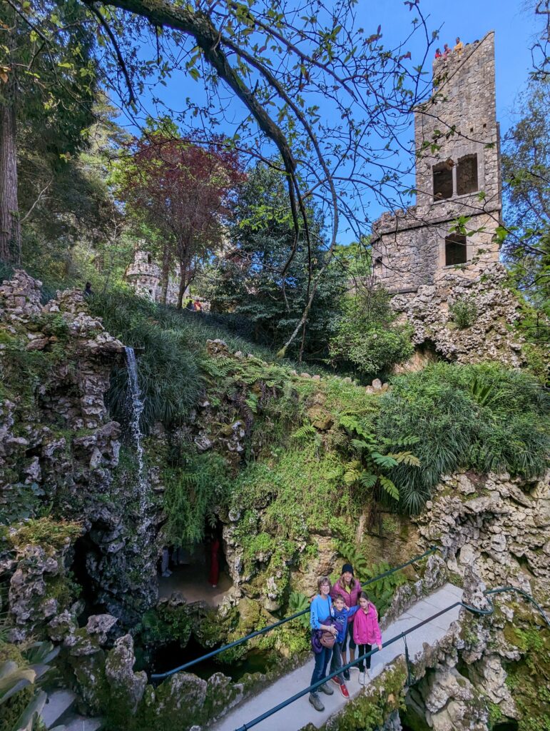 A family standing on a paved bridge with waterfall and tall stone tower at Quinta da Regaleira