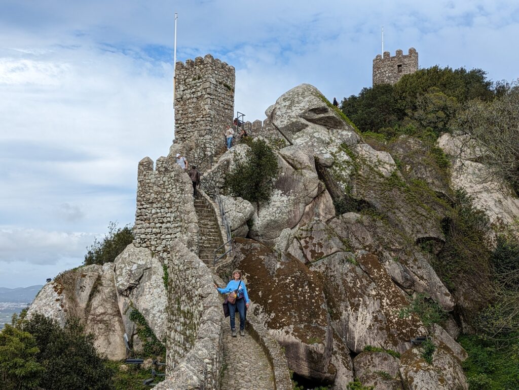 A woman standing at the bottom of an old stone staircase with castle walls and towers in the background