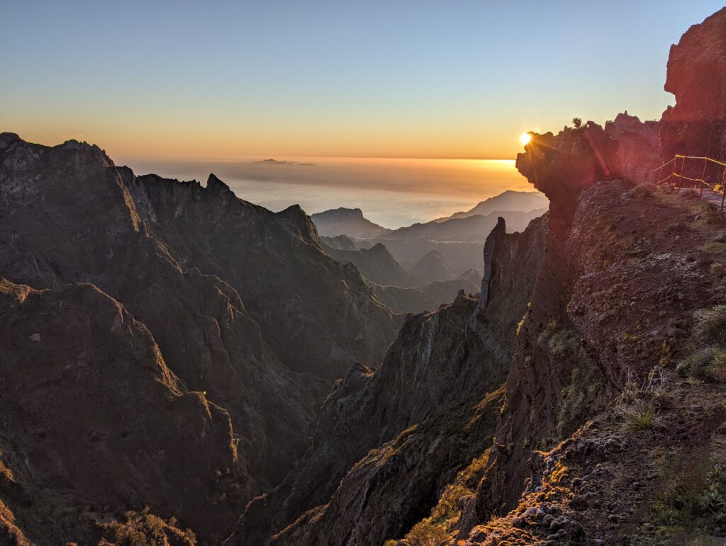 A jagged, shadowed mountain landscape with clouds and the sun rising in the background