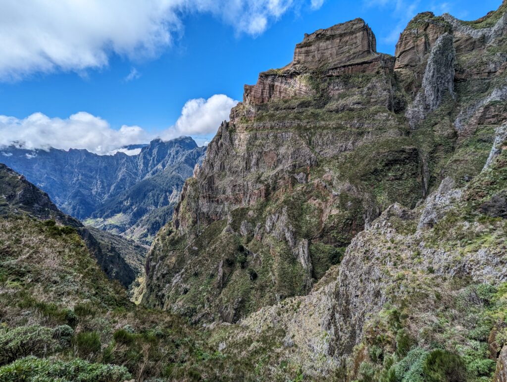 An imposing green mountain peak with other mountains in the background with blue skies and a few clouds