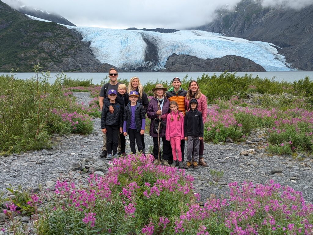 A group of people standing in front of a glacier with pink wildflowers in front of them