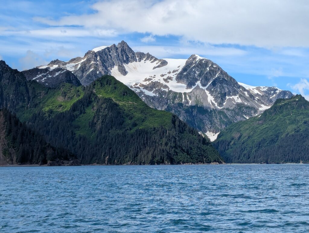 Snow-covered mountains surrounded by greenery with a sea in the foreground