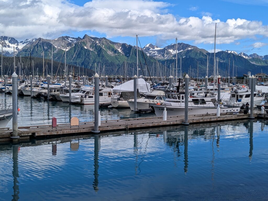 A dock and posts reflecting in the water with many boats and mountains in the background in Seward