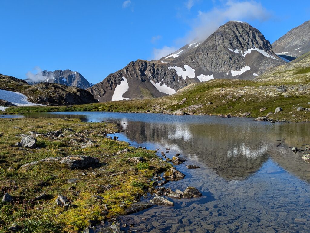 A rounded mountain reflected in a small pond with some grass and rocks in the foreground