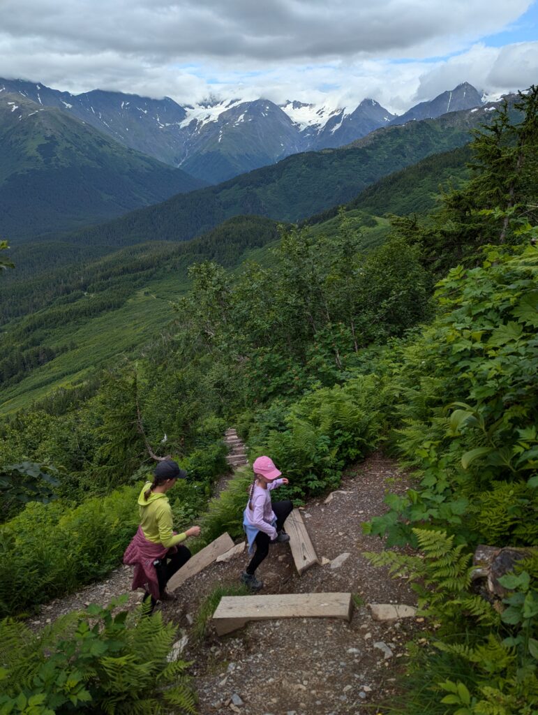 A mom and daughter climbing up a hiking trail with mountains and clouds in the background