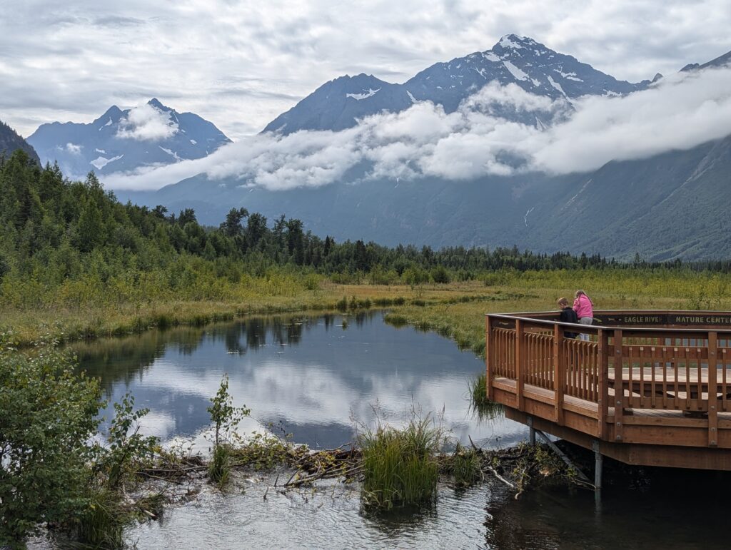 Two children leaning over a wooden railing on a pond with cloudy mountains at Eagle River Nature Center