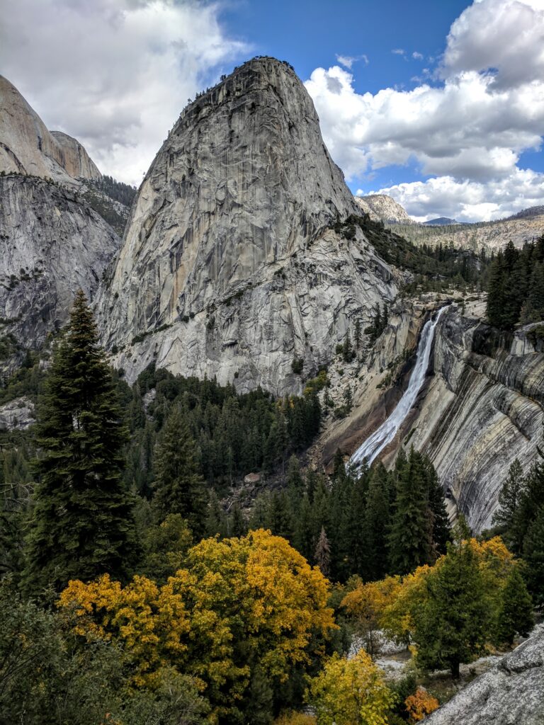 A waterfall cascading down a rock face with a giant rounded mountain behind it and yellow trees in the foreground