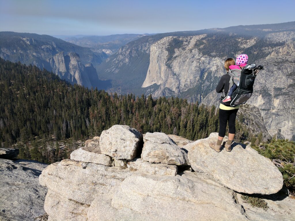 A woman with a young child in a backpack standing on a rock looking at a valley