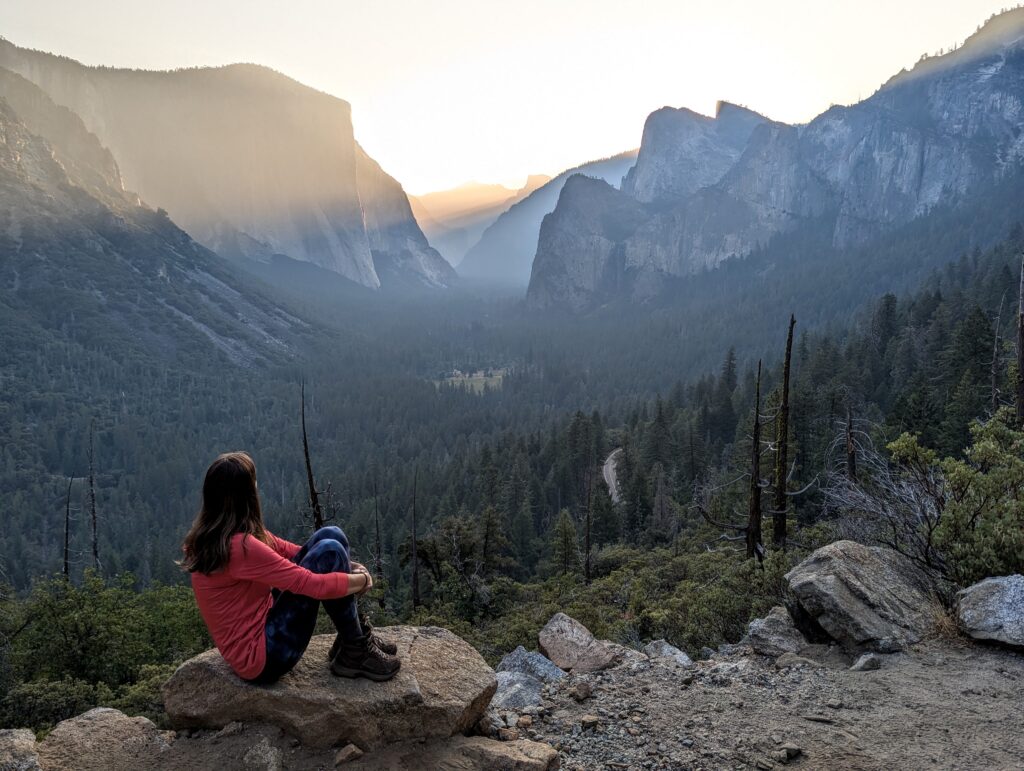 A woman sitting on a rock looking at the sun peaking through layers of mountains