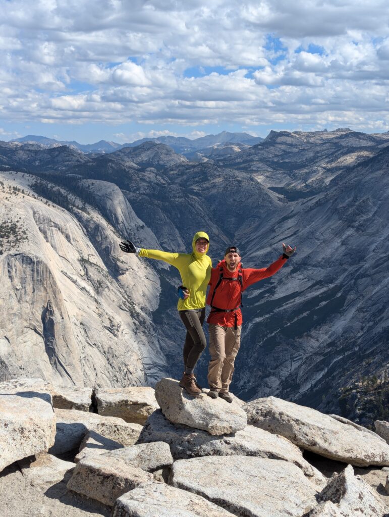 A couple standing towards the edge of a drop-off with a mountainous valley directly behind them