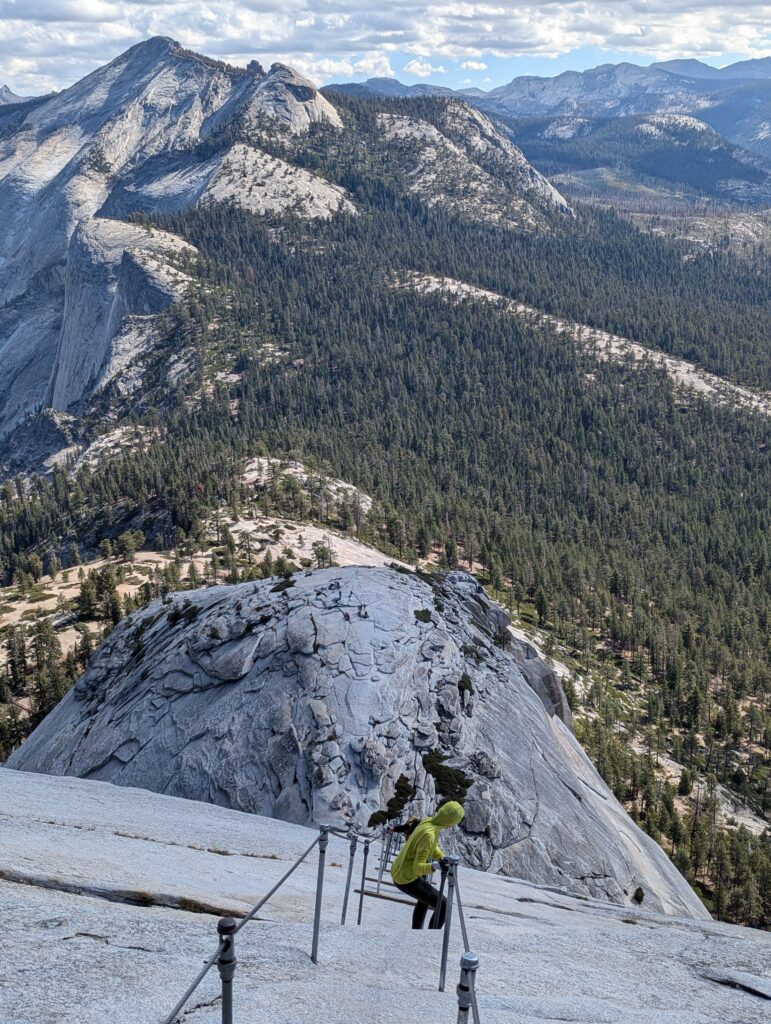 A hiker precariously descending a steep rock face with the help of cables on Half Dome