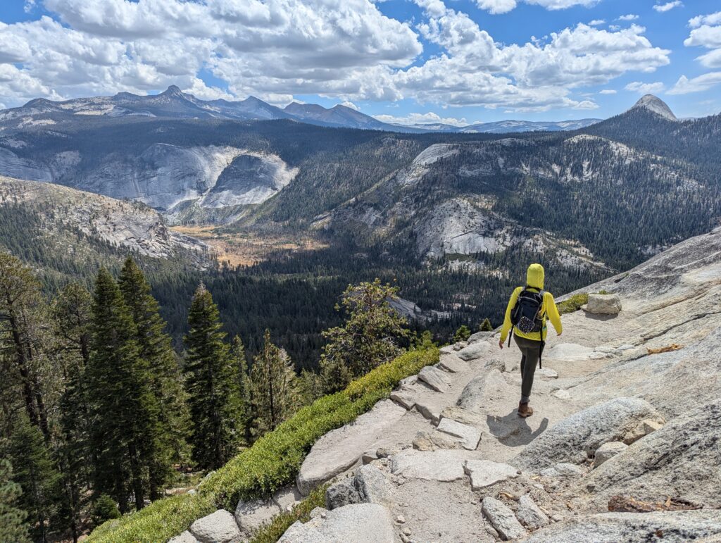 A person in a yellow shirt and backpack walking along a stone edge with trees and mountains