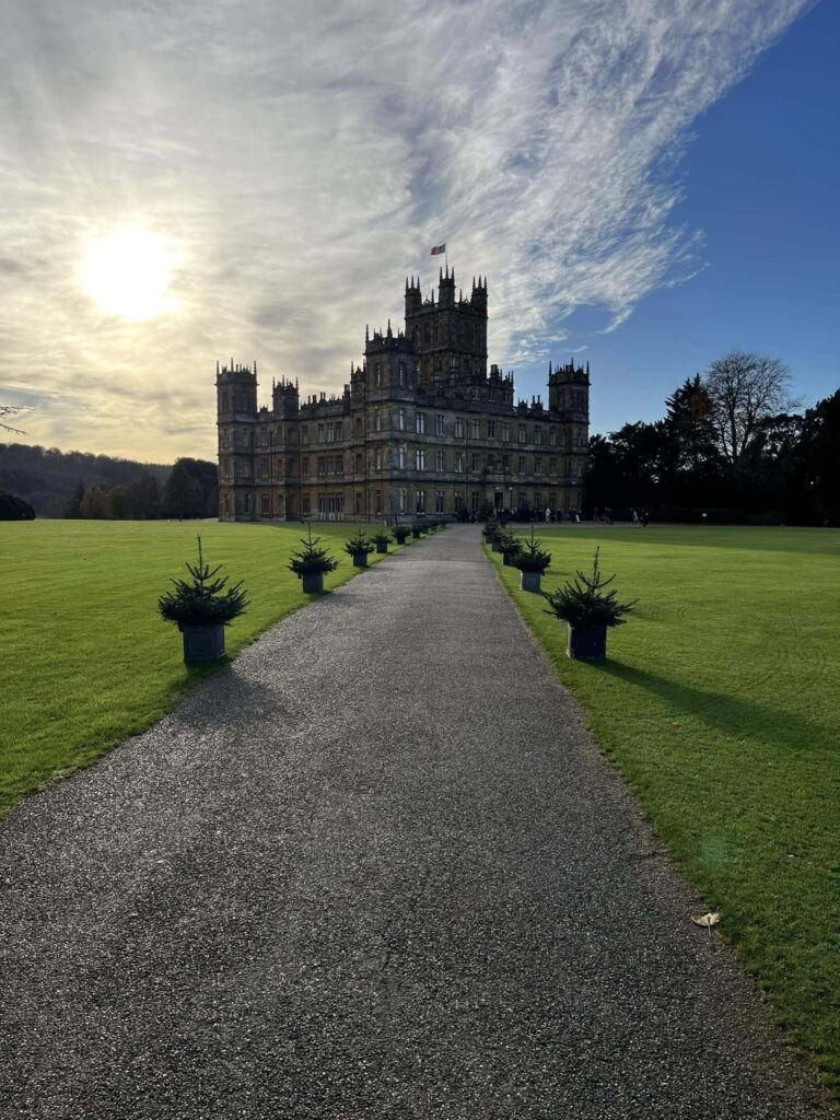 A stone path lined with small trees leading to Highclere Castle on a beautiful sunny and cloudy day