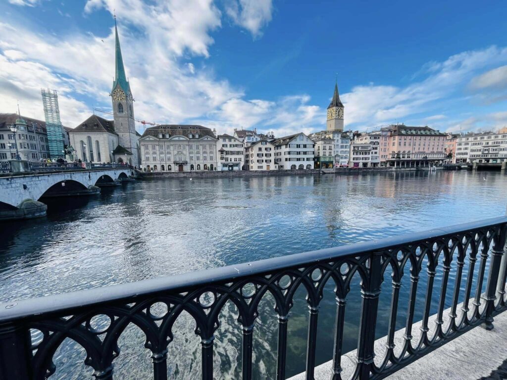 A railing leading to water with houses and towers across the way in Zurich